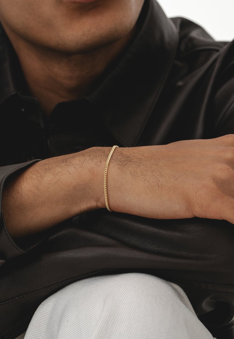 Goldene Kettenarmband an einem Handgelenk, mit einem verdrehten Design. Der Arm ruht auf einer schwarzen Lederoberfläche, mit einem teilweise sichtbaren weißen Stoff.