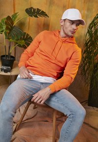 Sudadera naranja con cordón, camiseta blanca debajo, jeans de mezclilla claros. Sentado en una silla de madera, con una planta en maceta y una cámara vintage cerca.