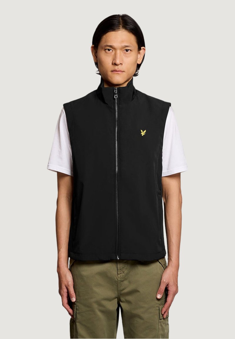 Uomo che indossa un gilet senza maniche nero con cerniera e logo giallo sopra una T-shirt bianca e pantaloni cargo oliva, in piedi contro uno sfondo semplice.