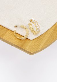 Orecchini a cerchio dorati con rhinestones trasparenti, caratterizzati da un design attorcigliato. Ambientati su un tessuto bianco testurizzato e una superficie in bambù.