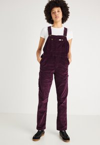 Salopette in velluto a coste viola con spalline regolabili, due tasche frontali e una vestibilità comoda, abbinata a una t-shirt bianca e sneakers nere.