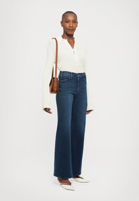 Donna che indossa una camicia bianca a maniche lunghe, jeans a vita alta e a gamba larga, scarpe bianche a punta e porta una borsa a tracolla marrone.