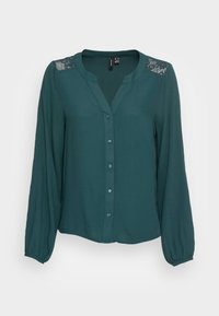 Blouse vert foncé avec un col en V, manches longues et accents en dentelle sur les épaules. Dotée d'une fermeture à boutons et d'un tissu lisse et léger.