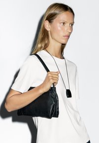 Borsa nera in pelle con forma strutturata, dal design semplice e manici corti, tenuta da una persona con una maglietta bianca.