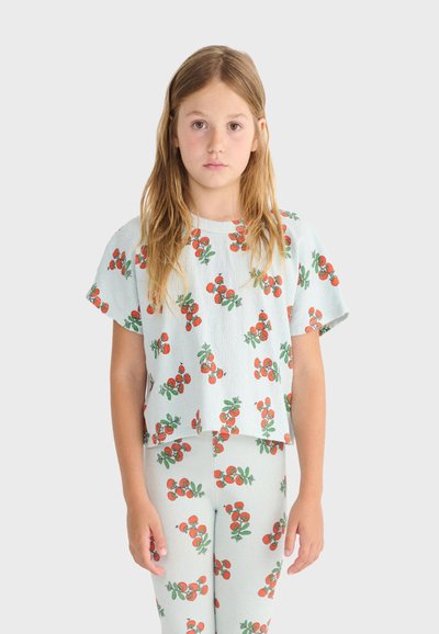 Jeune fille aux longs cheveux portant un haut et un legging bleu clair assortis avec des motifs de cerises rouges, debout face à l'avant sur un fond uni.