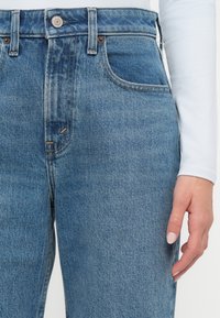 Κοντινό πλάνο ενός ατόμου που φοράει μπλε ψηλόμεσα τζιν και λευκό μακρυμάνικο τοπ, με ένα χέρι να στηρίζεται δίπλα στο μηρό.