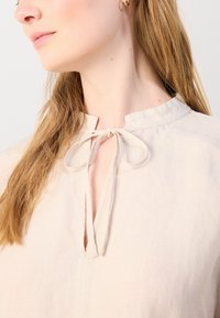 Světle béžový top s obšitým výstřihem a zavazováním v přední části. Tkanina vypadá jemně a má decentní strukturu.