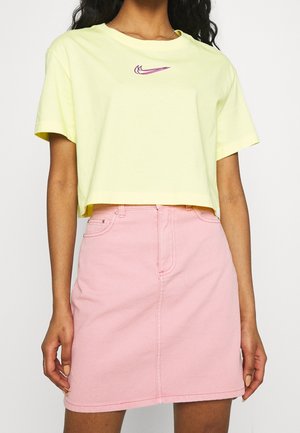 Femme portant un t-shirt Nike court jaune pâle avec un swoosh violet et une jupe en jean rose taille haute sur fond blanc.