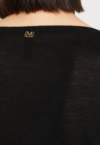 Czarny sweter z dzianiny z prążkowanym dekoltem, z złotym akcentem logo na plecach. Tekstura wydaje się miękka i lekko przezroczysta.