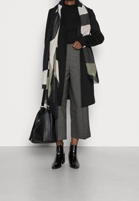 Czarny szalik w geometryczny wzór, czarny długi płaszcz, dopasowana czarna bluzka, szare kuloty, czarnebotki o teksturowanej powierzchni oraz czarna torebka.