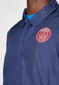 Dark-skinned person wearing a navy blue buttoned jacket with a red Paris Saint-Germain logo and red Nike swoosh on the chest.