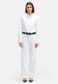 Biała koszula na długi rękaw z guzikami i kieszonką na piersi, zestawiona z białymi, wysokim stanem, szerokimi spodniami jeansowymi oraz czarnym paskiem. Czarne buty na obcasie z zaostrzonym noskiem.