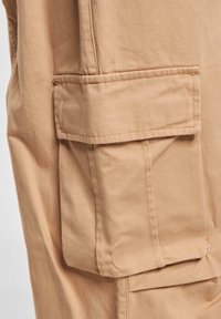 Beige cargo bukser med en lappelomme på låret, lavet af slidstærkt stof med synlige syninger. Glat tekstur, lige snit.