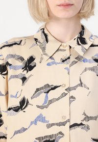 Beige button-up shirt featuring a printed pattern of black and blue abstract shapes. Textured fabric with a classic collar and pearl-button closure.