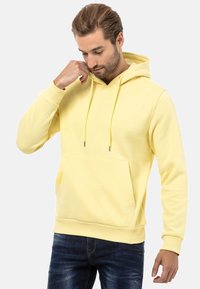 Gelbe Kapuzenjacke mit einer Fronttasche, strukturiertem Stoff und Kordeln. Die Bündchen und der Saum haben einen gerippten Abschluss. Dunkle Jeans getragen.
