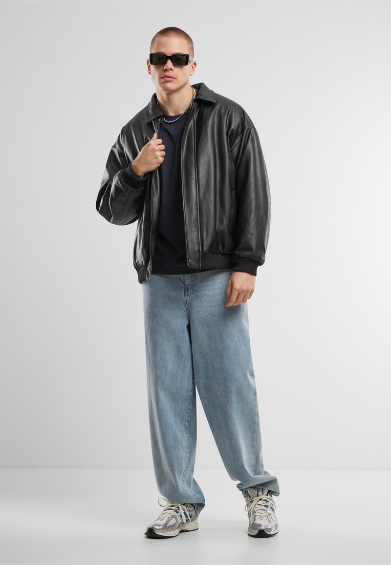 Jachetă de piele neagră tip bomber, pantaloni din denimovești de mari dimensiuni, adidași gri și albi și ochelari de soare negri. Modelul stă în fața unui fundal gri.