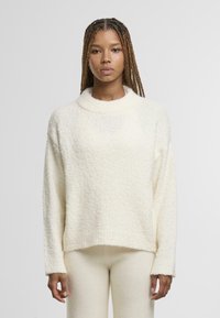 Žena s opletenými vlasy v bílé texturované dlouhé mikině a odpovídajících kalhotách, stojící na jednobarevném světlém pozadí.