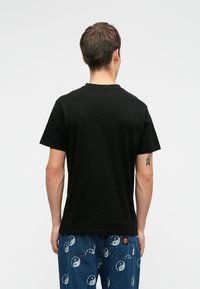 Άνδρας που στέκεται με πλάτη προς την κάμερα, φορώντας μαύρο μπλουζάκι και μπλε τζιν, εκτυπωμένα με λευκά σύμβολα γιν-γιάνγκ και μία μικρή κόκκινη στάμπα.