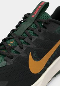 Black and green athletic shoe with a textured mesh upper, black laces, orange Nike Swoosh, and a red tag at the heel.