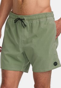 Shorts de bain verts avec ceinture élastique, cordon de serrage noir, poches latérales et un patch logo circulaire en bas. Texture lisse.