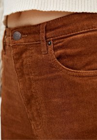Gros plan sur la poche et la ceinture d'un pantalon en velours côtelé marron avec un bouton métallique, porté avec un pull en maille beige clair.