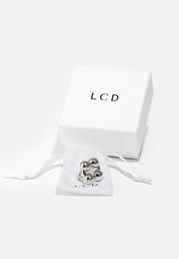 Anillo de metal plateado con acentos esféricos presentado en una bolsa de tela blanca con cordón, junto a una caja blanca etiquetada "LCD" en negro.
