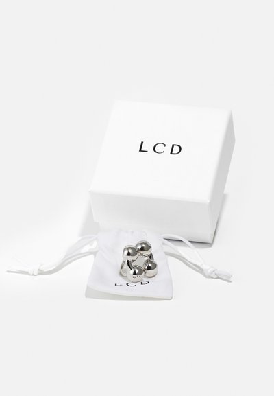 Zilveren metalen ring met bolvormige accenten, gepresenteerd in een witte trekkoordzak, naast een witte doos met het opschrift "LCD" in zwart.