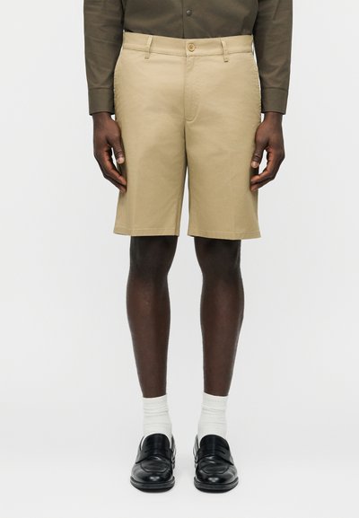 Homme portant un short beige jusqu'aux genoux, des chaussettes blanches, des mocassins en cuir noir et une chemise marron à manches longues, debout devant un fond uni.