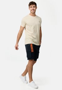 Beige kortærmet t-shirt med knaplukning ved halsen og lille logo, parret med mørkeblå cargo shorts og hvide sneakers.