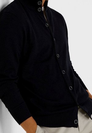 Homme portant un cardigan noir boutonné sur une chemise blanche, les mains dans les poches d'un pantalon clair, sur fond blanc.