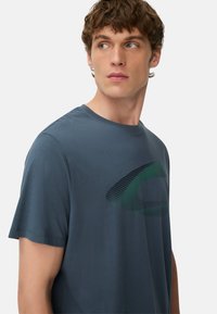 Ung mand med kort, krøllet brunt hår, iført en mørkeblå T-shirt med et grønt og sort abstrakt design, kigger til siden.