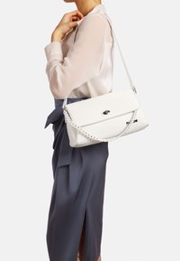 Borsa a tracolla in pelle bianca con texture, chiusura a patta e dettagli in metallo argentato, posizionata su uno sfondo neutro.