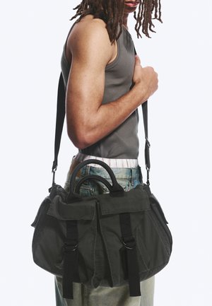Person with shoulder-length dreadlocks wears gray tank top and jeans, carrying large black canvas shoulder bag with buckled pockets.