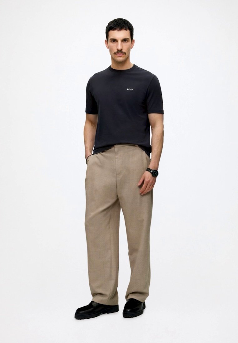 Homme avec moustache portant un t-shirt noir BOSS, pantalon beige à jambes larges, montre noire et chaussures noires, debout les mains dans les poches.