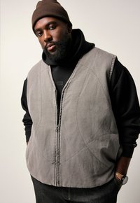 Grau gepolsterte Weste mit einem Reißverschluss, die mit geschwungenen Nähmustern und einem hohen Kragen versehen ist, getragen über einem schwarzen Hoodie. Braune gestrickte Mütze getragen.