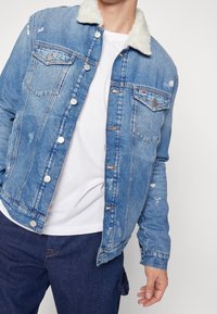 Blå denimjacka med vit fuskpälskrage, slitna detaljer, knäppning fram, två bröstfickor och avslappnad passform. Buren över en vit T-shirt.