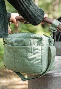 Töltött zöld szövet táska vállpánttal és felső fogantyúval, elülső zsebbel és puha, párnázott textúrával.