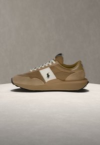 Basket beige et marron clair avec des détails en mesh, des lacets blancs et un panneau blanc avec un logo de joueur de polo noir, présentée sur une surface claire.