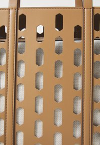 Tan perforeren nep leren tas met hexagonale uitsparingen die een witte, gestructureerde linnen binnenkant blootleggen, voorzien van stevige handvaten.