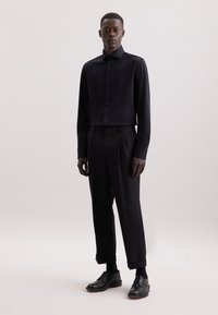 Maglietta nera a maniche lunghe con colletto e bottoni, abbinata a pantaloni neri a vita alta con pieghe e scarpe eleganti nere. Texture liscia.