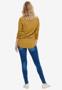 Gul långärmad tröja med rundad nederkant, i kombination med blå skinny jeans och vita sneakers. Håret stylat i lösa vågor.