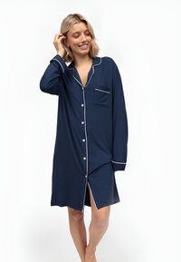 Chemise de nuit bleu marine avec passepoils blancs, boutons sur le devant, une poche poitrine, des manches longues et une fente en bas. Tissu doux.
