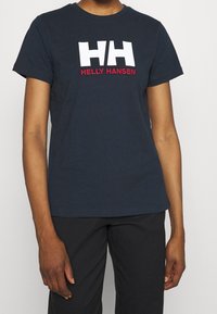 Námořnická modrá bavlněná tričko s bílým logem "HH" a červeným textem "HELLY HANSEN". Krátké rukávy a standardní kulatý výstřih.