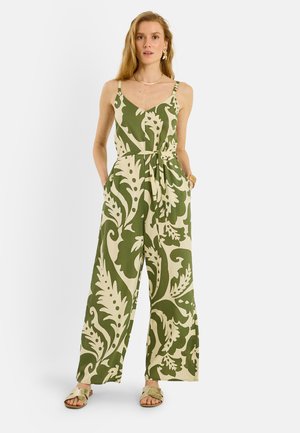 Mujer con un mono sin mangas verde y blanco con estampado floral, de piernas anchas y sandalias, de pie con las manos en los bolsillos sobre un fondo blanco.