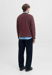 Maglione a maglia color mogano con collo rotondo indossato sopra una camicia di denim azzurro chiaro, abbinato a pantaloni scamosciati scuri e scarpe marroni, visto da dietro.