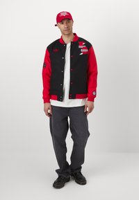 Giacca varsity nera e rossa con bottoni a pressione, decorata con toppe e ricami, abbinata a una camicia bianca, jeans scuri e scarpe nere.