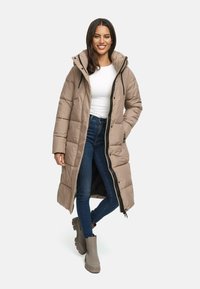 Lang, beige puffer-jakke med lynlås foran og hætte, med en quiltet design; parret med mørke jeans og grå ankelstøvler.
