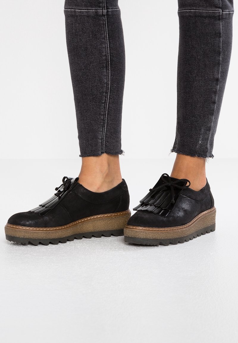 Chaussures à plateforme en daim noir avec semelle épaisse et texturée, ornées de franges décoratives et d'une fermeture à lacets. Portées avec un jean skinny foncé.
