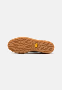 Semelle de chaussure avec motif de bande de roulement en zigzag en caoutchouc marron clair et un petit logo hexagonal jaune près de la zone de la voûte plantaire.
