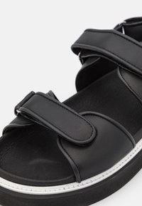 Mustat sandaalit, joissa on säädettävät Velcro-vöitä, sileä synteettinen päällinen, pehmustettu sisävuori ja paksu valkoinen ommeltu ulkopohja.